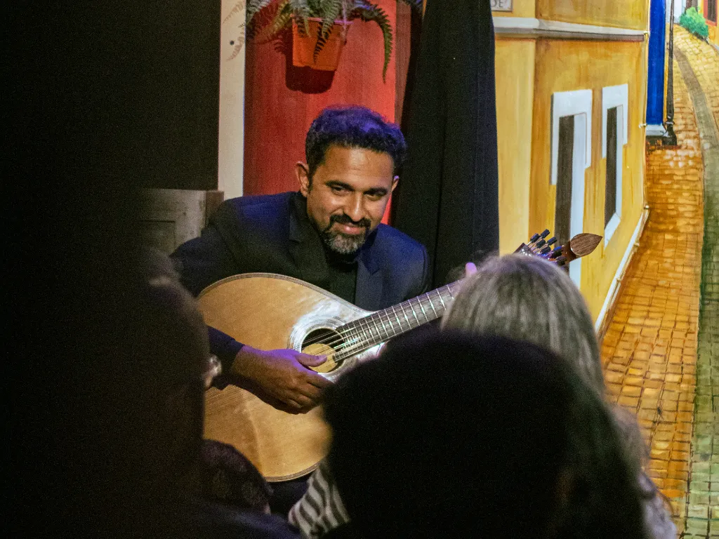 Smiling man in a dark blazer plays an acoustic guitar on a small stage, facing an audience.
