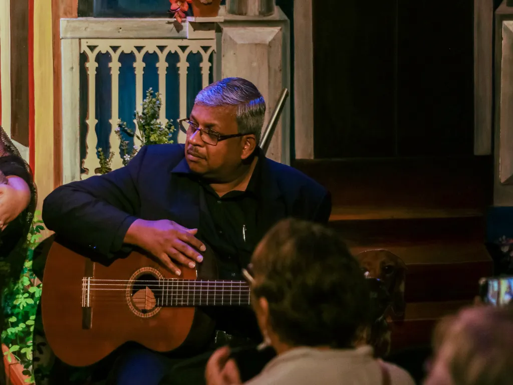 Middle-aged man with glasses playing an acoustic guitar at a small indoor venue, seated near a decorative railing.