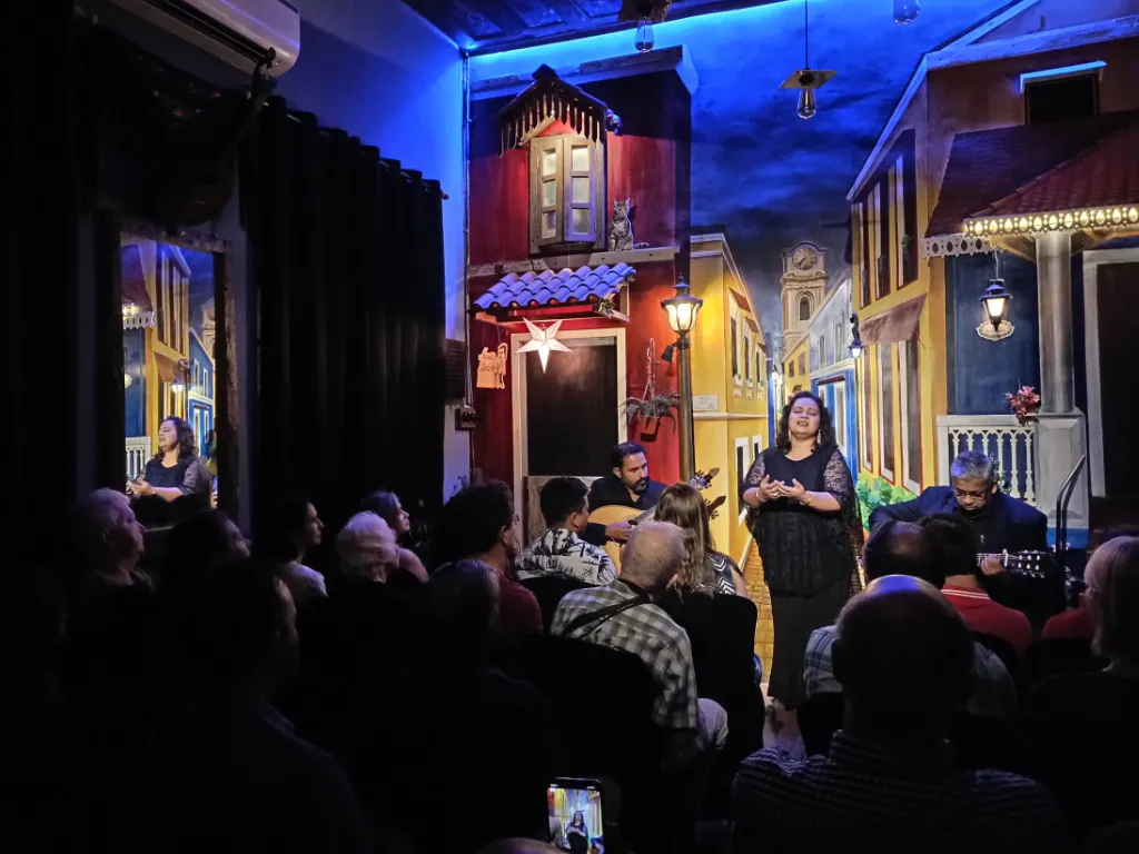 Female singer in a black dress performs center stage with guitar and pianist beside her, set against a colorful painted street backdrop, audience in front.