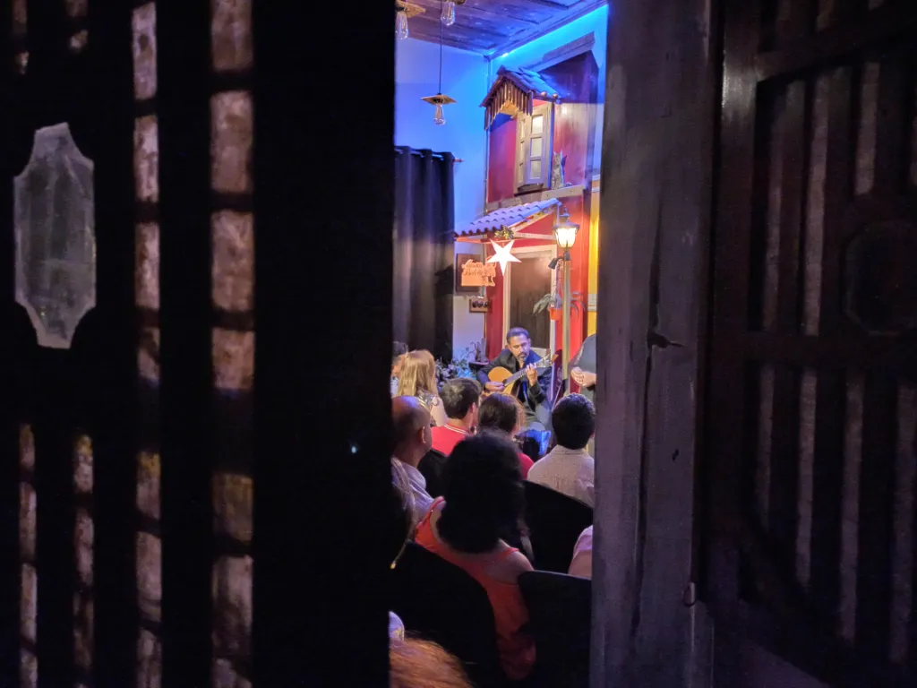 Guitarist performs on a small colorful stage inside a cozy venue, viewed through a dark doorway with seated audience in front.