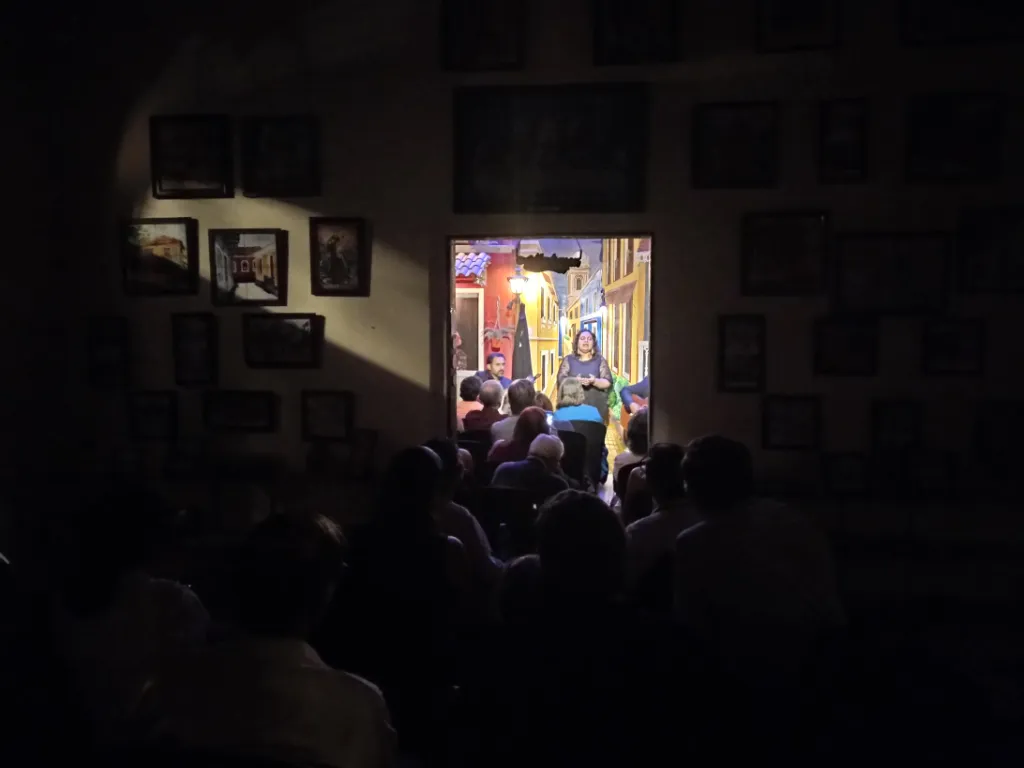 Dim room with audience seated in darkness, a performer speaks from a brightly lit doorway framed by colorful murals.