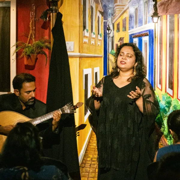 Woman in a black shawl sings with eyes closed as a guitarist plays beside a colorful painted backdrop.