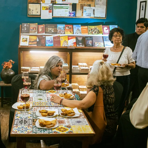 Group of four adults seated at a tiled cafe table with pastries and red wine, bookshelves and displays in the background.