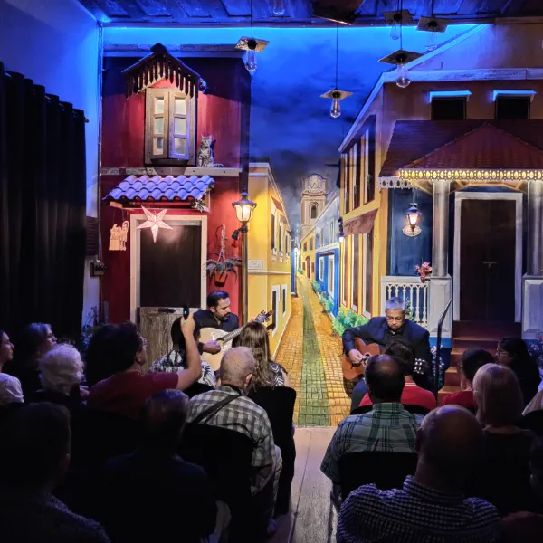 Two guitarists perform on a colorful stage lined with a painted town street; audience seated in front, blue overhead lights.