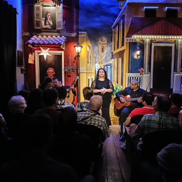 Singer stands center stage with two guitarists seated beside her in a colorful street-set venue, performing to a seated audience.