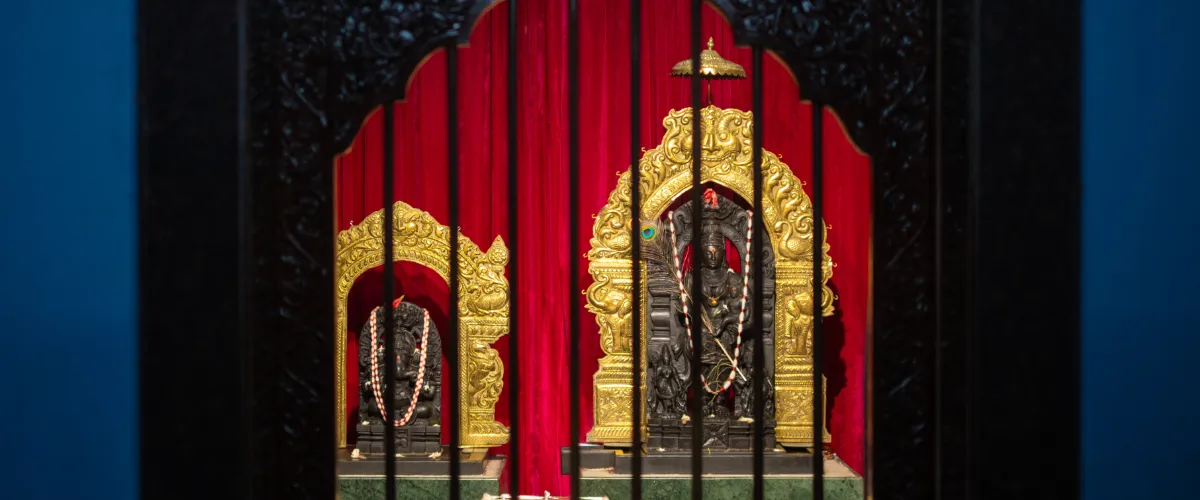 A shrine with two ornate golden arches housing dark statues, red velvet backdrop, and braided garlands in front barrier bars.