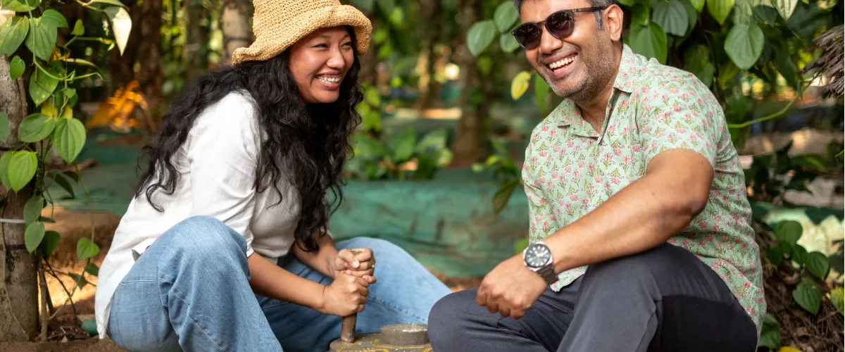 Two adults gardening together, smiling as they dig in a sunlit yard with leafy plants around them.