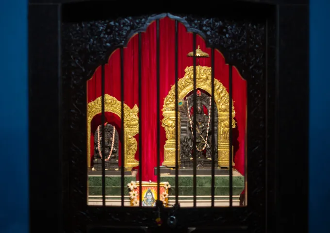 Hindu shrine behind vertical bars with gold ornate arches and red drapery, small framed image and flower arrangement on a pedestal at the base.