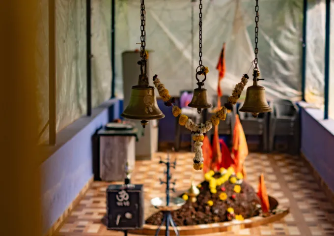 Bronze bells hanging above a small Hindu shrine on a tiled floor, with a flower garland and orange flags in the background.