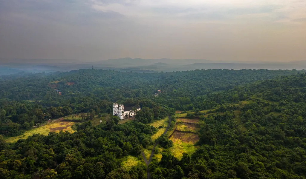 Aerial view of a white building nestled in dense green forest and rolling hills, with fields nearby under a hazy sky, eyes on nature and solitude.