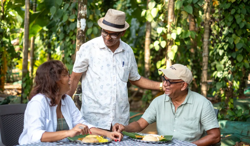 Three friends share a meal at an outdoor garden table, smiling and chatting together under leafy trees.