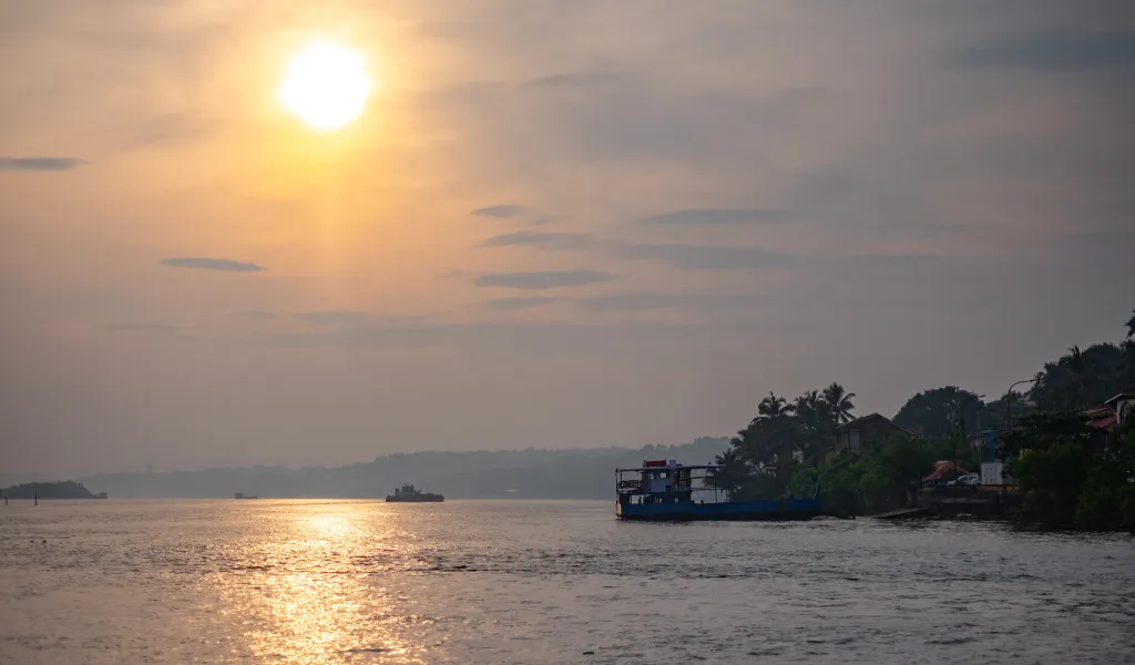 Sun setting over a calm river with a boat near the shore and palm trees on the right side.