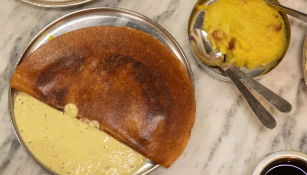 Masala dosa on a metal plate with coconut chutney on the side and potato curry nearby on a marble table